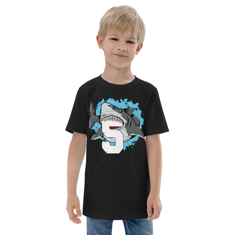 May include: Black short-sleeved t-shirt with a graphic of a shark biting a number 5. The shark is white and gray with a blue background.