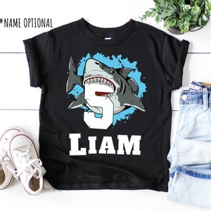 May include: Black t-shirt with a blue and white graphic of a shark with the number 5 and the name Liam.