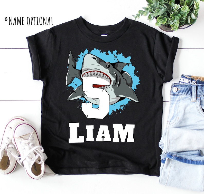 May include: Black t-shirt with a blue and white graphic of a shark with the number 5 and the name Liam.