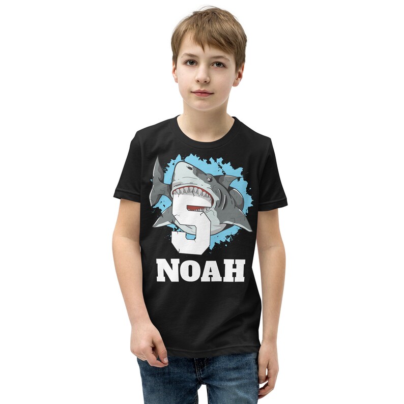 May include: Black t-shirt with a blue and white graphic of a shark with the number 5 inside its mouth. The text "NOAH" is printed below the shark.