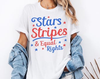 May include: A white short-sleeve t-shirt featuring the blue and red text Stars Stripes & Equal Rights. The design includes small blue and red stars scattered around the lettering. The shirt is worn with a blue denim jacket and denim shorts.