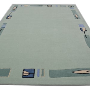 Op de afbeelding: Een lichtgroen tapijt met een blauwe en crèmekleurige gepatenteerde rand. De rand heeft gestileerde bladeren en geometrische vormen.