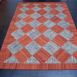 Puede incluir: Una alfombra rectangular con un patrón de diamantes en tonos naranja y gris. La alfombra tiene un borde naranja y gris.