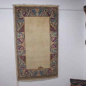 May include: A beige rug with a blue and purple floral border. The rug has a signature in the center.