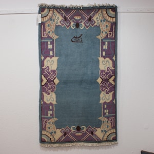 May include: A light blue rug with a purple and cream border. The rug has a traditional Chinese design with floral motifs. The rug is hanging on a white wall. The rug has a label that says "HAN'S".
