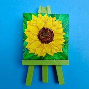 Puede incluir: Una peque&ntilde;a pintura sobre lienzo de un girasol en un caballete verde. El girasol es amarillo con un centro marr&oacute;n y hojas verdes.