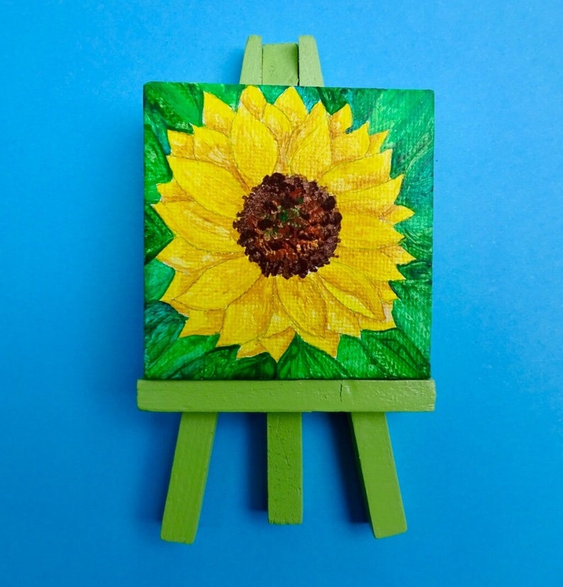 Puede incluir: Una peque&ntilde;a pintura sobre lienzo de un girasol en un caballete verde. El girasol es amarillo con un centro marr&oacute;n y hojas verdes.