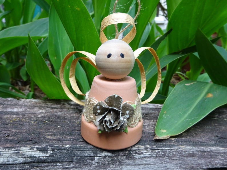 K&ouml;nnte beinhalten: Ein kleiner Engel-Anh&auml;nger aus einem Terrakotta-Topf mit einem Holzkopf, goldenen Papierfl&uuml;geln und einem Juteblumen-Akzent.