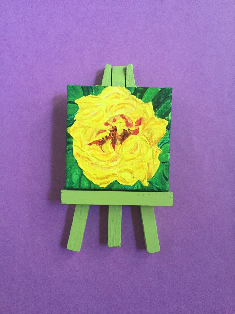 Puede incluir: Una peque&ntilde;a pintura sobre lienzo de una flor amarilla en un caballete verde. La flor est&aacute; pintada en un estilo estilizado e impresionista.