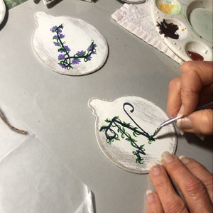 May include: A person is painting a white wooden ornament with a black and green floral design. The ornament is round and has a loop at the top for hanging. The person is using a small paintbrush to apply the paint.
