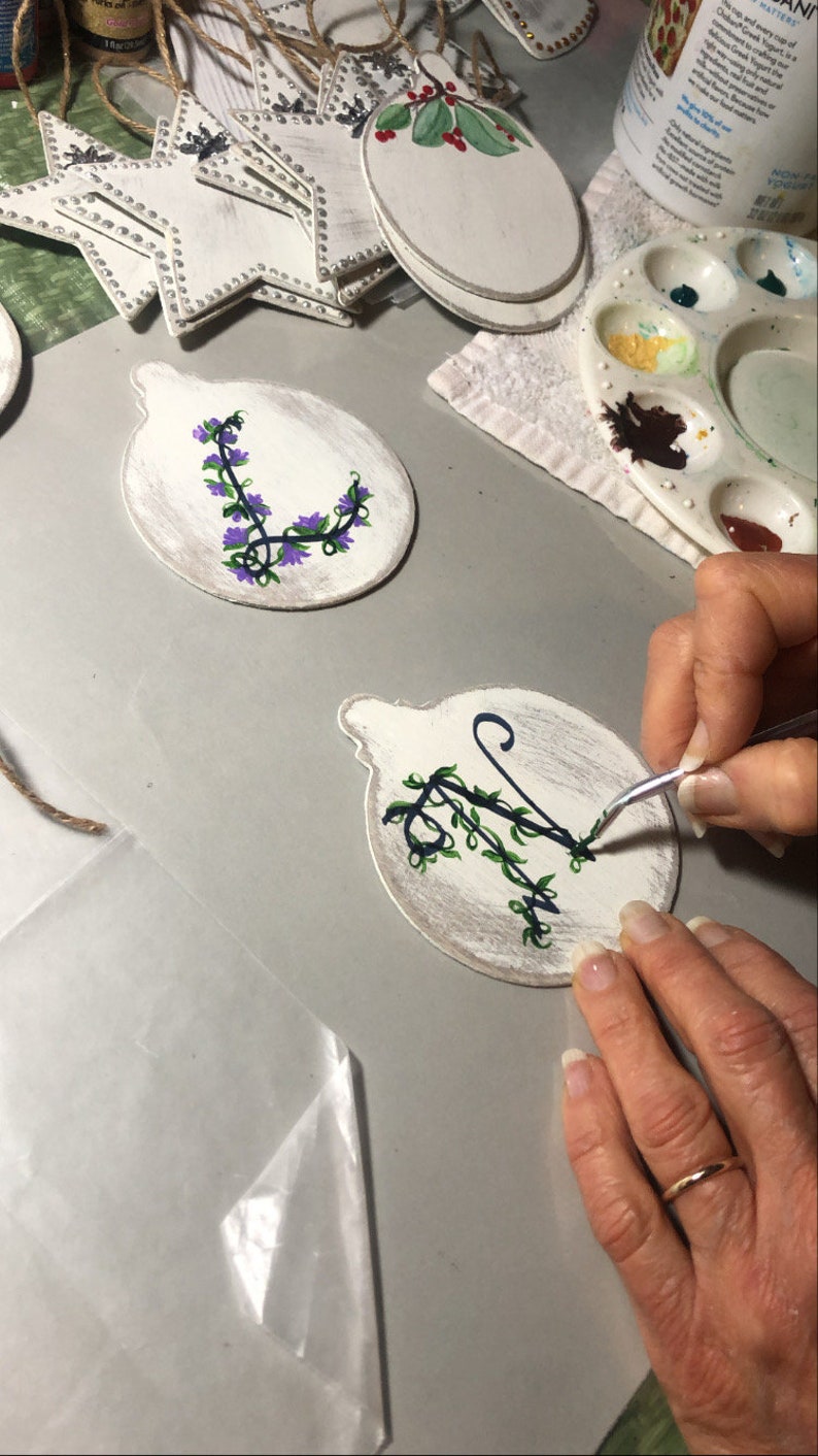 May include: A person is painting a white wooden ornament with a black and green floral design. The ornament is round and has a loop at the top for hanging. The person is using a small paintbrush to apply the paint.