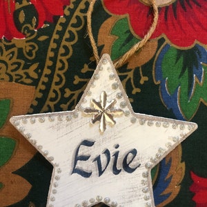 May include: White wooden star ornament with a silver snowflake and the name "Evie" written in black paint. The star is decorated with silver glitter around the edges.