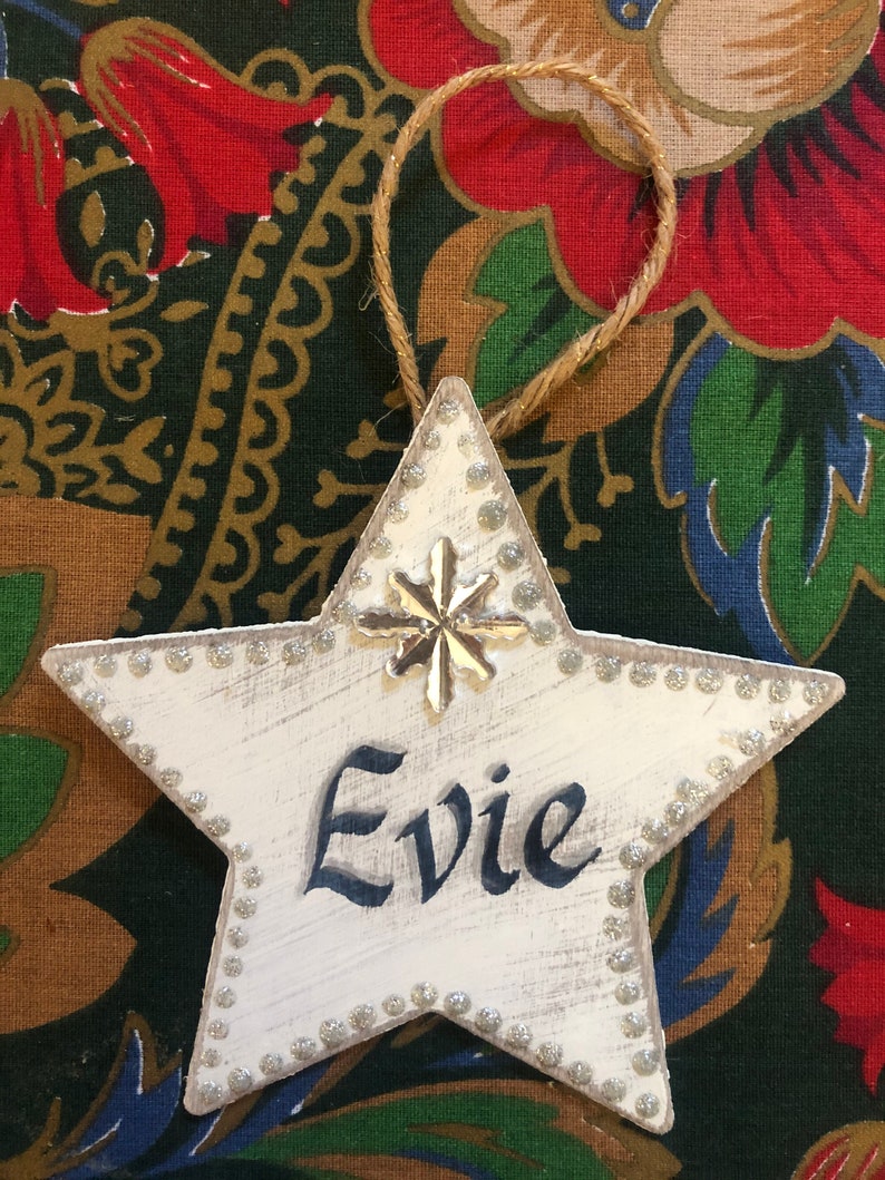May include: White wooden star ornament with a silver snowflake and the name "Evie" written in black paint. The star is decorated with silver glitter around the edges.