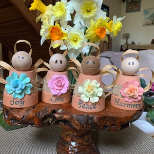 K&ouml;nnte beinhalten: Vier kleine Terrakotta-Blument&ouml;pfe, die als Engel mit Fl&uuml;geln dekoriert sind, jeder mit einer andersfarbigen Blume und den Worten "Freude", "Liebe", "Frieden" und "Harmonie" auf der Vorderseite.