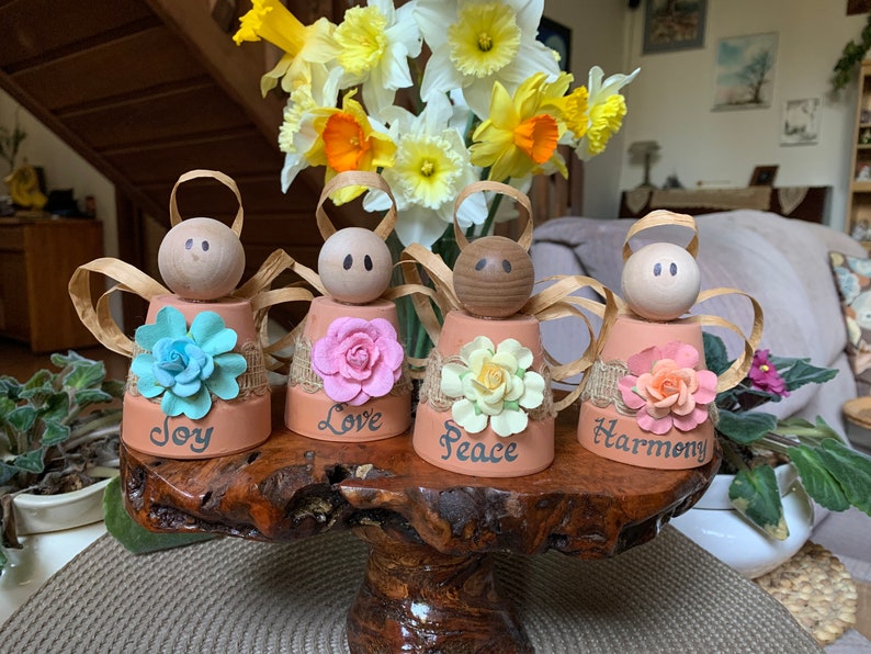K&ouml;nnte beinhalten: Vier kleine Terrakotta-Blument&ouml;pfe, die als Engel mit Fl&uuml;geln dekoriert sind, jeder mit einer andersfarbigen Blume und den Worten "Freude", "Liebe", "Frieden" und "Harmonie" auf der Vorderseite.