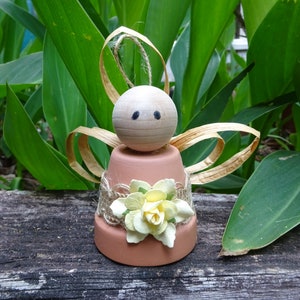 K&ouml;nnte beinhalten: Eine kleine Engelfigur aus einem Terrakotta-Topf mit einem Holzkopf und Strohfluegeln. Der Engel ist mit einem Jutesband und gelben Papierblumen verziert.
