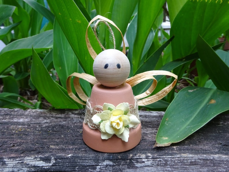 K&ouml;nnte beinhalten: Eine kleine Engelfigur aus einem Terrakotta-Topf mit einem Holzkopf und Strohfluegeln. Der Engel ist mit einem Jutesband und gelben Papierblumen verziert.