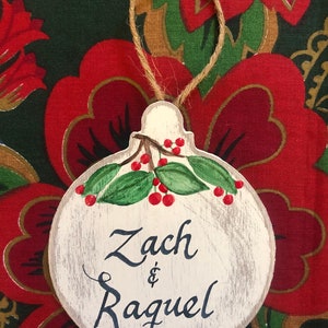 May include: A white wooden ornament shaped like a Christmas tree bauble with a green holly sprig and red berries painted on it. The ornament is decorated with the names "Zach & Raquel" written in black cursive.