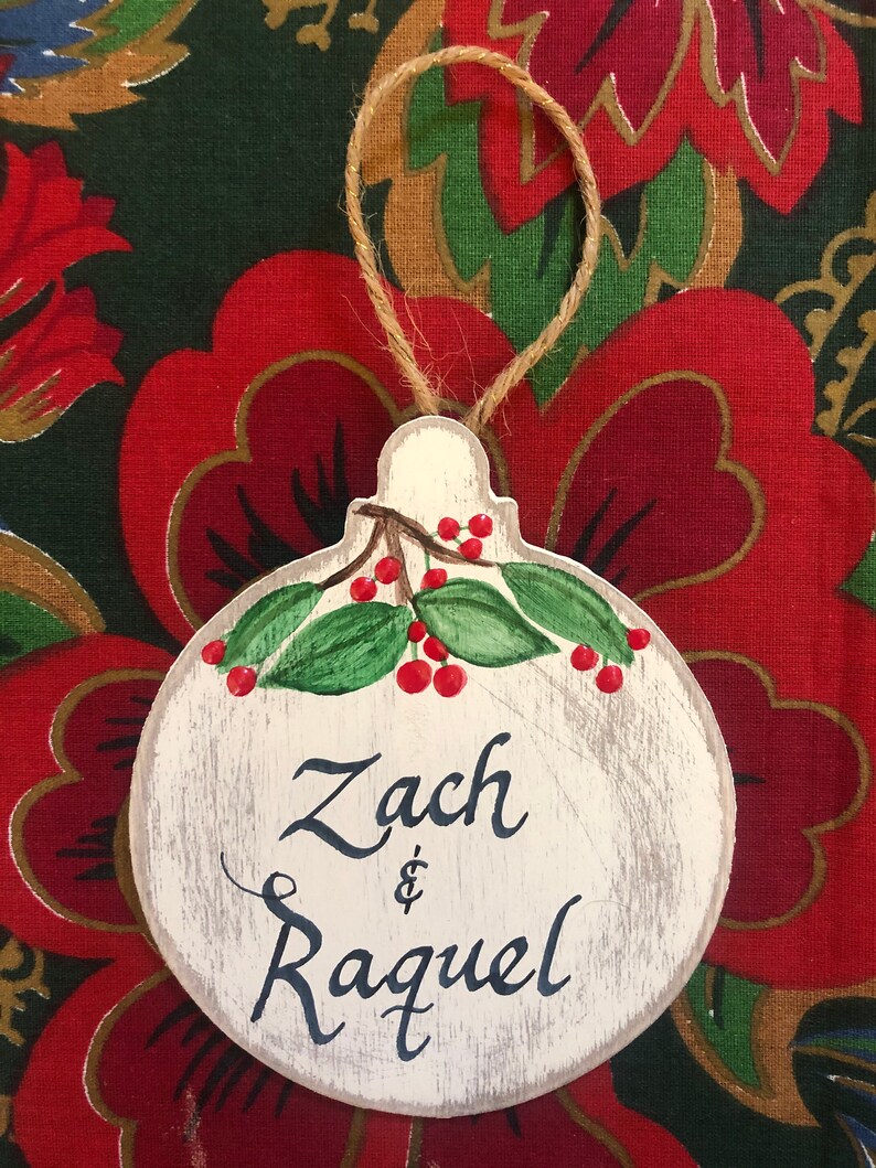 May include: A white wooden ornament shaped like a Christmas tree bauble with a green holly sprig and red berries painted on it. The ornament is decorated with the names "Zach & Raquel" written in black cursive.