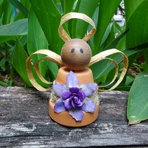 K&ouml;nnte beinhalten: Eine kleine Engelfigur aus einem Terrakotta-Topf mit einem braunen Holzkopf und Strohfluegeln. Der Topf ist mit einer lila Blume und Jute verziert.