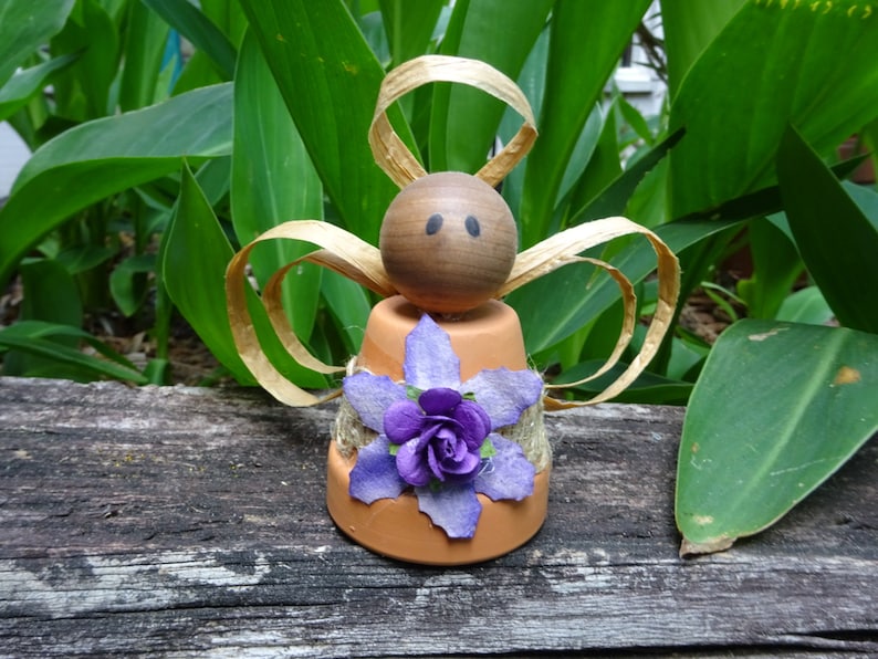 K&ouml;nnte beinhalten: Eine kleine Engelfigur aus einem Terrakotta-Topf mit einem braunen Holzkopf und Strohfluegeln. Der Topf ist mit einer lila Blume und Jute verziert.