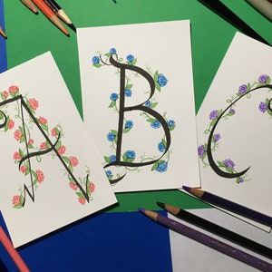 May include: Three cards with the letters A, B, and C written in black ink with a vine and flower design around each letter. The flowers are pink, blue, and purple.