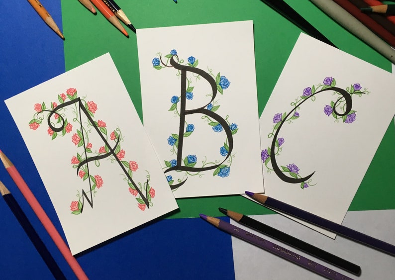 May include: Three cards with the letters A, B, and C written in black ink with a vine and flower design around each letter. The flowers are pink, blue, and purple.