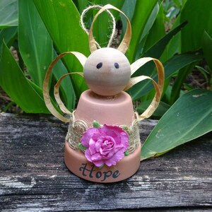K&ouml;nnte beinhalten: Eine kleine Engelfigur aus einem Terrakotta-Topf mit einem Holzkopf, braunen Bindfadenfl&uuml;geln und einer rosa Stoffblume. Der Topf ist mit Sackleinen verziert und das Wort "Hope" steht auf der Vorderseite.
