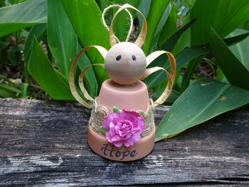 K&ouml;nnte beinhalten: Eine kleine Engelfigur aus einem Terrakotta-Topf mit einem Holzkopf, braunen Bindfadenfl&uuml;geln und einer rosa Stoffblume. Der Topf ist mit Sackleinen verziert und das Wort "Hope" steht auf der Vorderseite.