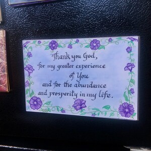 May include: A white card with a blue background and a purple floral border. The card has a handwritten message that reads "Thank you God, for my greater experience of You and for the abundance and prosperity in my life."