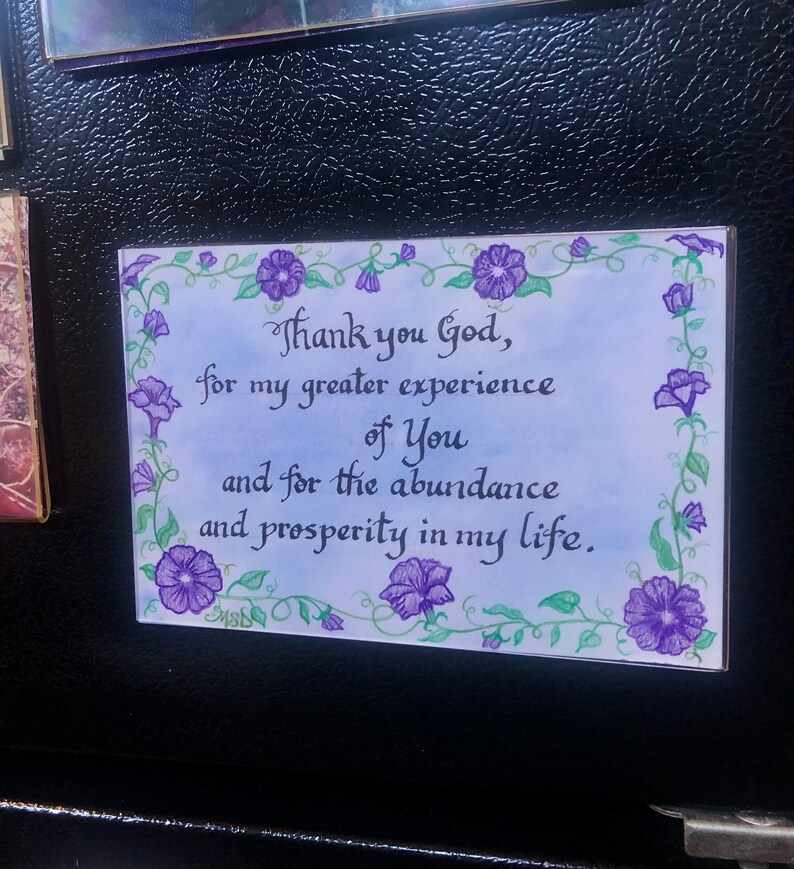 May include: A white card with a blue background and a purple floral border. The card has a handwritten message that reads "Thank you God, for my greater experience of You and for the abundance and prosperity in my life."