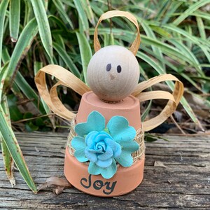 K&ouml;nnte beinhalten: Eine kleine Engelfigur aus einem Terrakotta-Topf mit einem hellbraunen Holzkopf, einer blauen Blumenverzierung und dem Wort "Freude" auf der Basis.