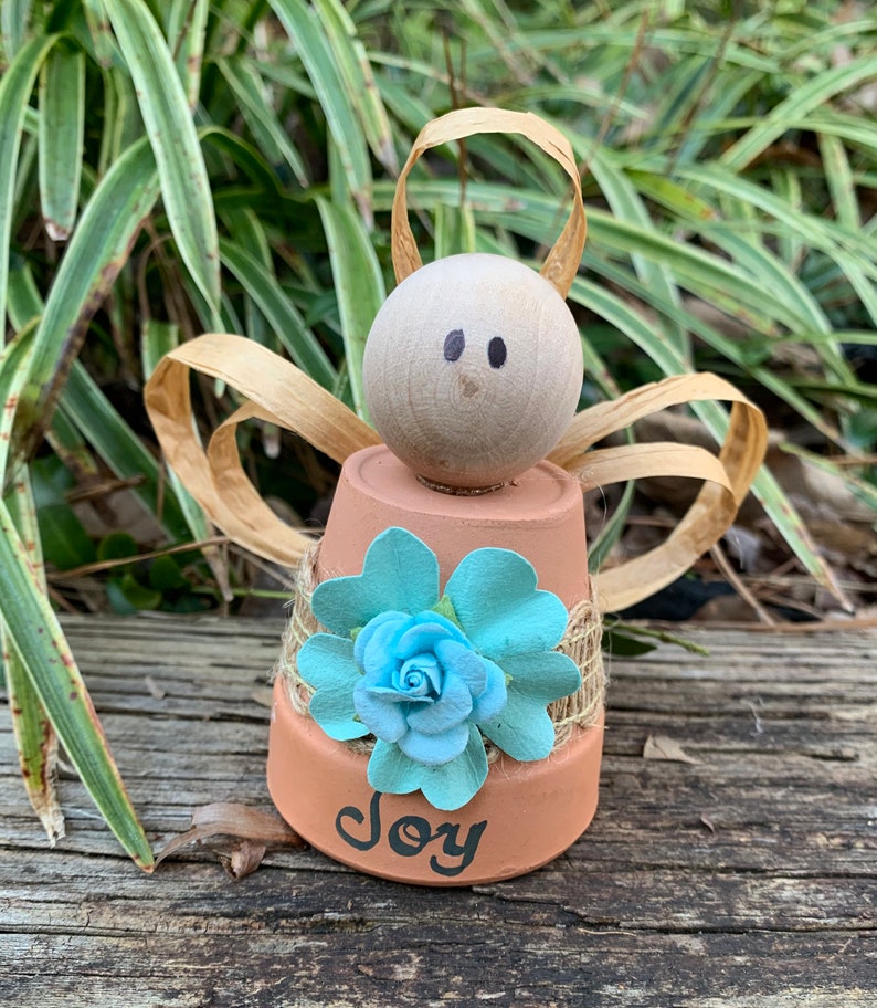K&ouml;nnte beinhalten: Eine kleine Engelfigur aus einem Terrakotta-Topf mit einem hellbraunen Holzkopf, einer blauen Blumenverzierung und dem Wort "Freude" auf der Basis.