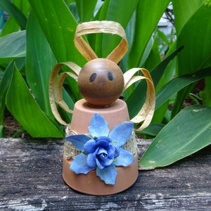 K&ouml;nnte beinhalten: Eine kleine Engelfigur aus einem Terrakotta-Topf mit einem braunen Holzkopf, einer blauen Blumenverzierung und goldenen Schleifenfl&uuml;geln.