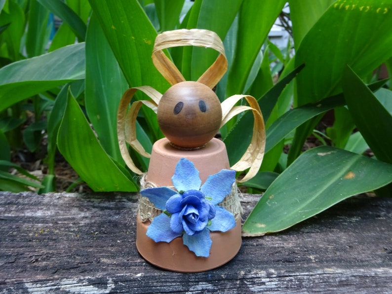 K&ouml;nnte beinhalten: Eine kleine Engelfigur aus einem Terrakotta-Topf mit einem braunen Holzkopf, einer blauen Blumenverzierung und goldenen Schleifenfl&uuml;geln.
