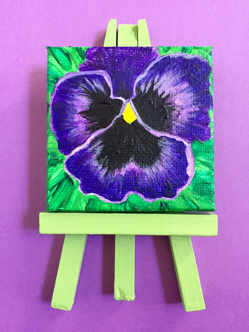 Puede incluir: Una flor de pensamiento pintada de morado y verde en un peque&ntilde;o caballete verde.