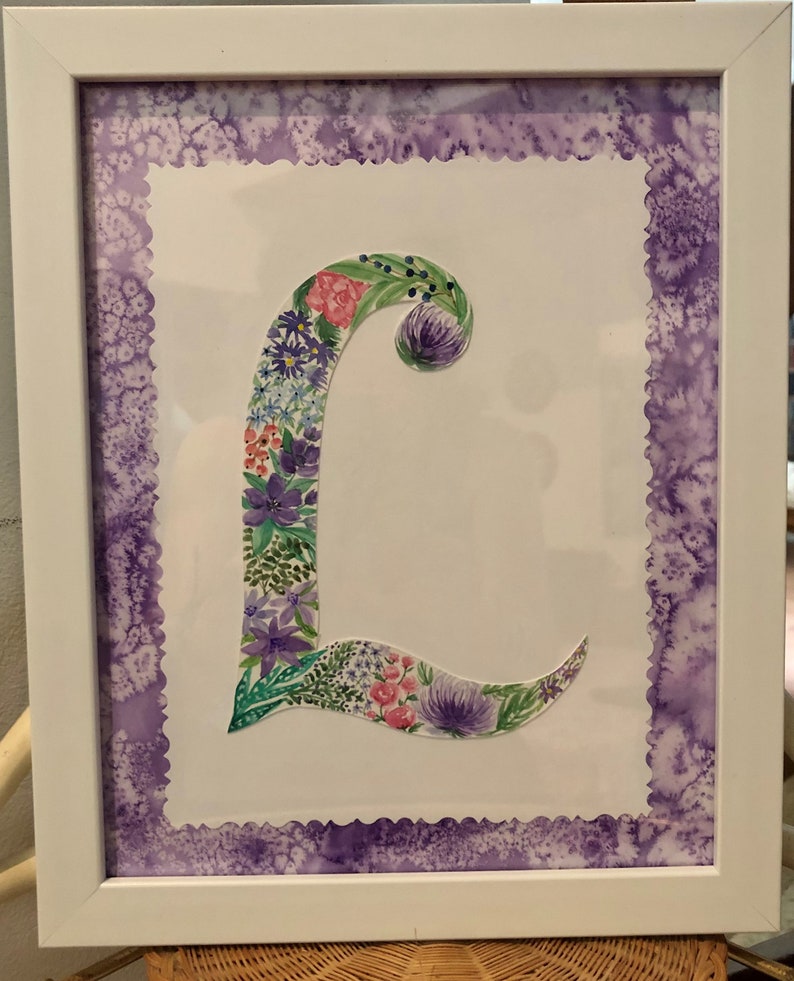 May include: A framed watercolor painting of the letter L, decorated with purple, pink, and green flowers. The frame has a purple scalloped border.