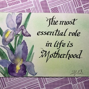 May include: A watercolor painting of purple irises with green leaves on a light green background. The text "The most essential role in life is Motherhood." is written in black cursive script. The painting is signed "M.S."