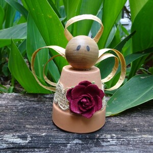 K&ouml;nnte beinhalten: Eine kleine Engelfigur aus einem Terrakotta-Topf mit einem Holzkopf, braunen Augen und einer burgunderfarbenen Stoffrose. Die Fl&uuml;gel des Engels sind aus goldenem Band.