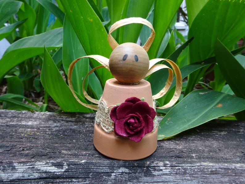 K&ouml;nnte beinhalten: Eine kleine Engelfigur aus einem Terrakotta-Topf mit einem Holzkopf, braunen Augen und einer burgunderfarbenen Stoffrose. Die Fl&uuml;gel des Engels sind aus goldenem Band.