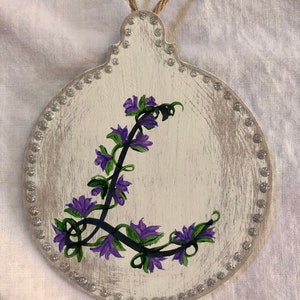 May include: A white wooden ornament with a silver glitter border. The ornament is painted with a purple flower design.