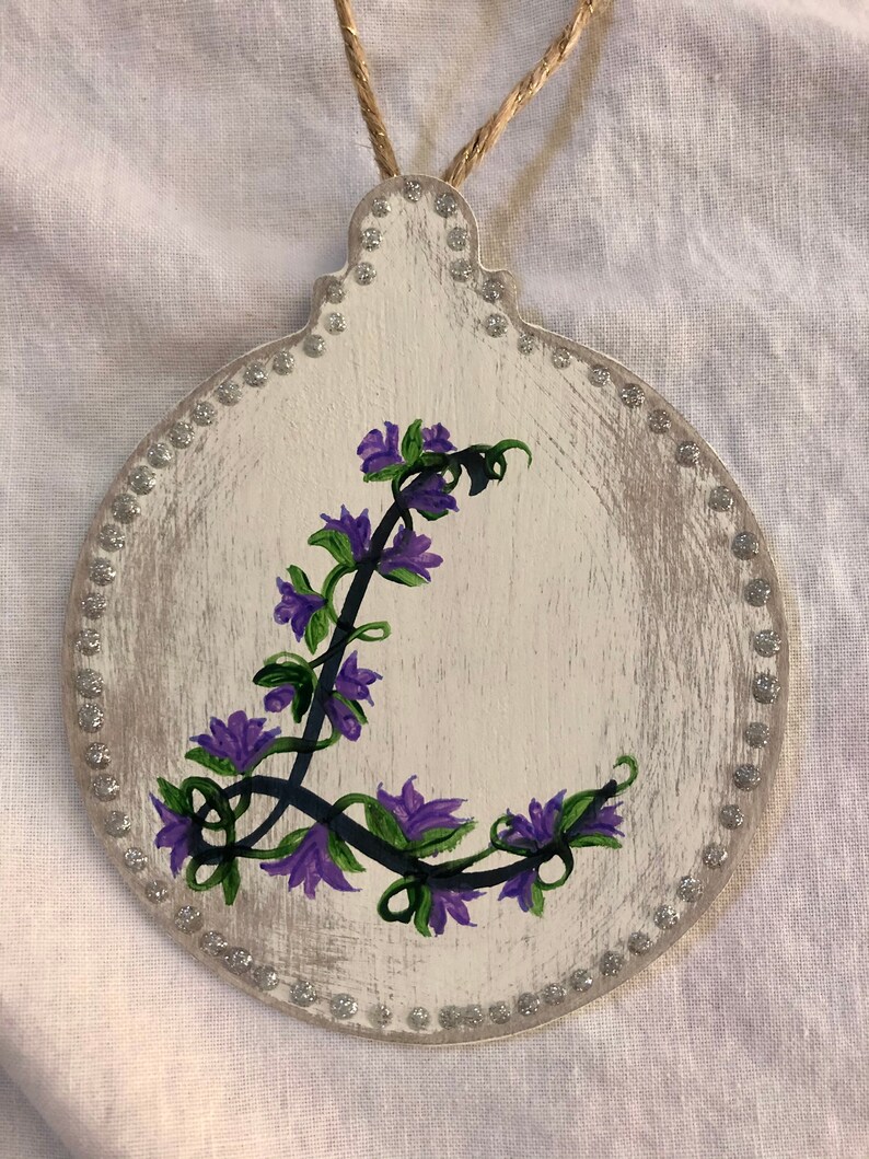 May include: A white wooden ornament with a silver glitter border. The ornament is painted with a purple flower design.