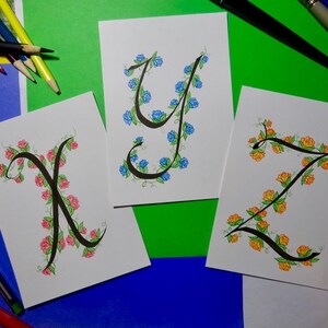 May include: Three cards with hand-lettered calligraphy of the letters X, Y, and Z. Each letter is surrounded by a vine of flowers. The X is surrounded by pink flowers, the Y is surrounded by blue flowers, and the Z is surrounded by yellow flowers.