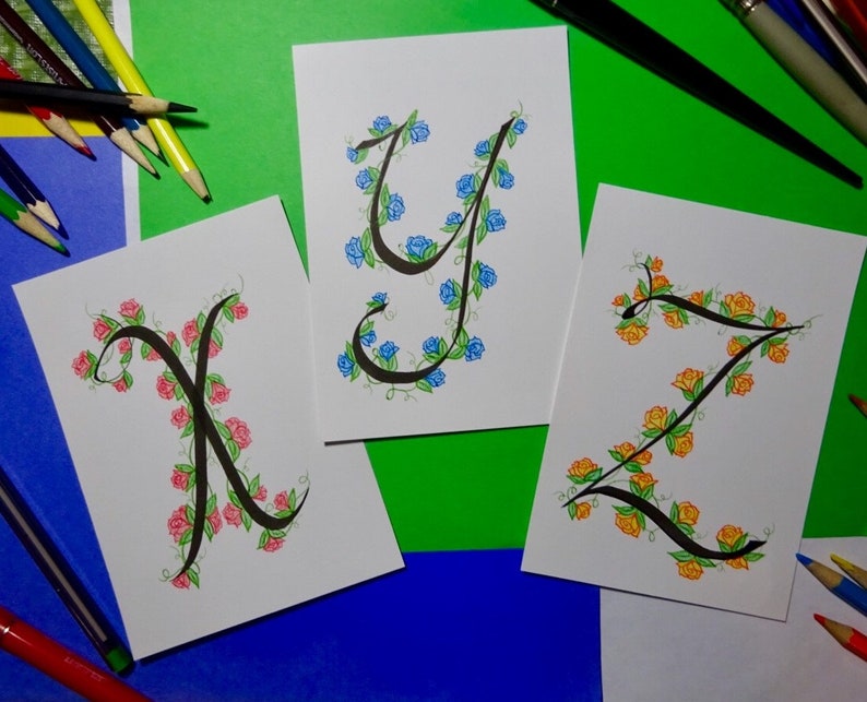 May include: Three cards with hand-lettered calligraphy of the letters X, Y, and Z. Each letter is surrounded by a vine of flowers. The X is surrounded by pink flowers, the Y is surrounded by blue flowers, and the Z is surrounded by yellow flowers.