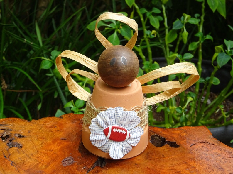 Peut inclure: Un pot de fleurs en terre cuite avec une boule en bois brun sur le dessus, d&eacute;cor&eacute; d'un ruban en toile de jute brun et d'une fleur en papier avec un ballon de football rouge et blanc au centre. Le pot est entour&eacute; d'un ruban dor&eacute;.