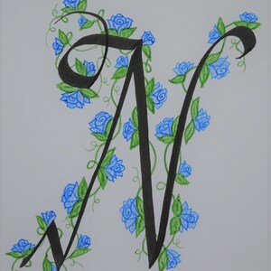May include: A black calligraphic letter N with blue and green floral embellishments. The letter is surrounded by blue roses and green leaves.