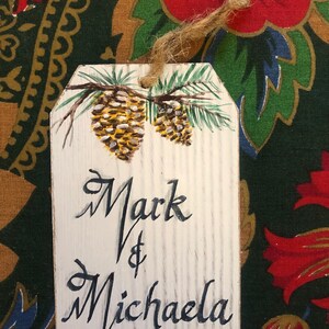 May include: White wooden gift tag with a painted pine branch and pine cones. The tag is tied with twine and has the text "Mark & Michaela" written in black cursive.