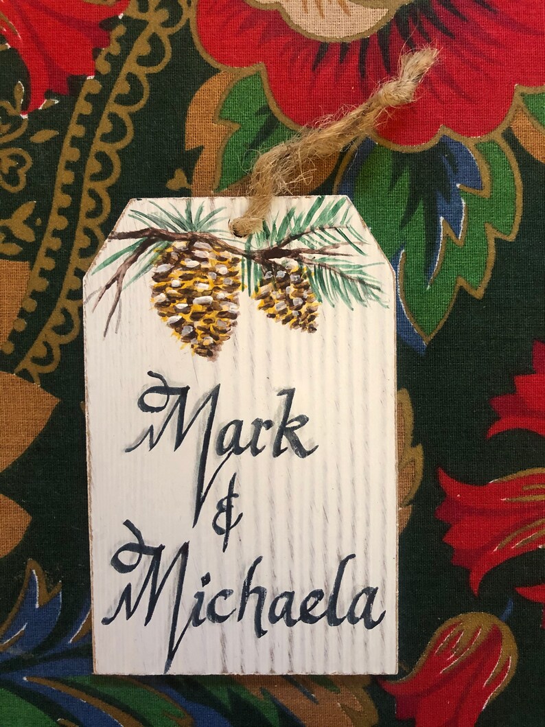 May include: White wooden gift tag with a painted pine branch and pine cones. The tag is tied with twine and has the text "Mark & Michaela" written in black cursive.