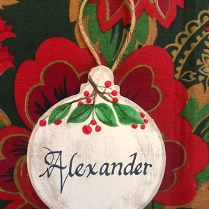 May include: A white wooden Christmas ornament with a red berry sprig and the name "Alexander" written in black cursive.