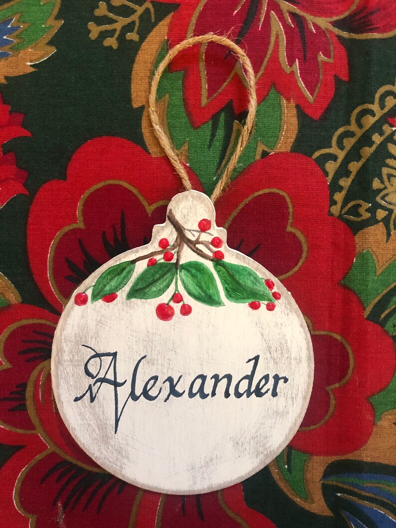 May include: A white wooden Christmas ornament with a red berry sprig and the name "Alexander" written in black cursive.